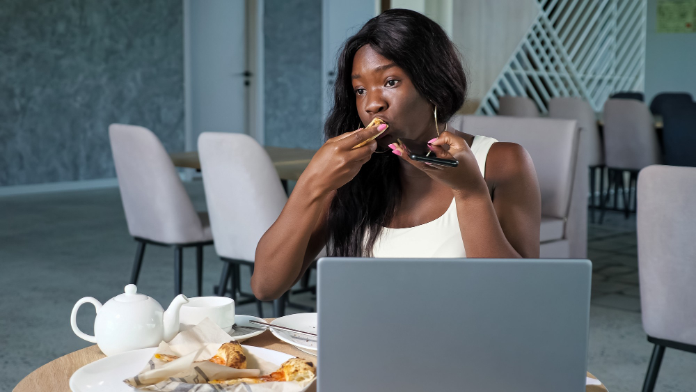 Young long haired black woman eats pizza talks using loudspeaker mode on phone near open modern laptop on table in light contemporary cafe