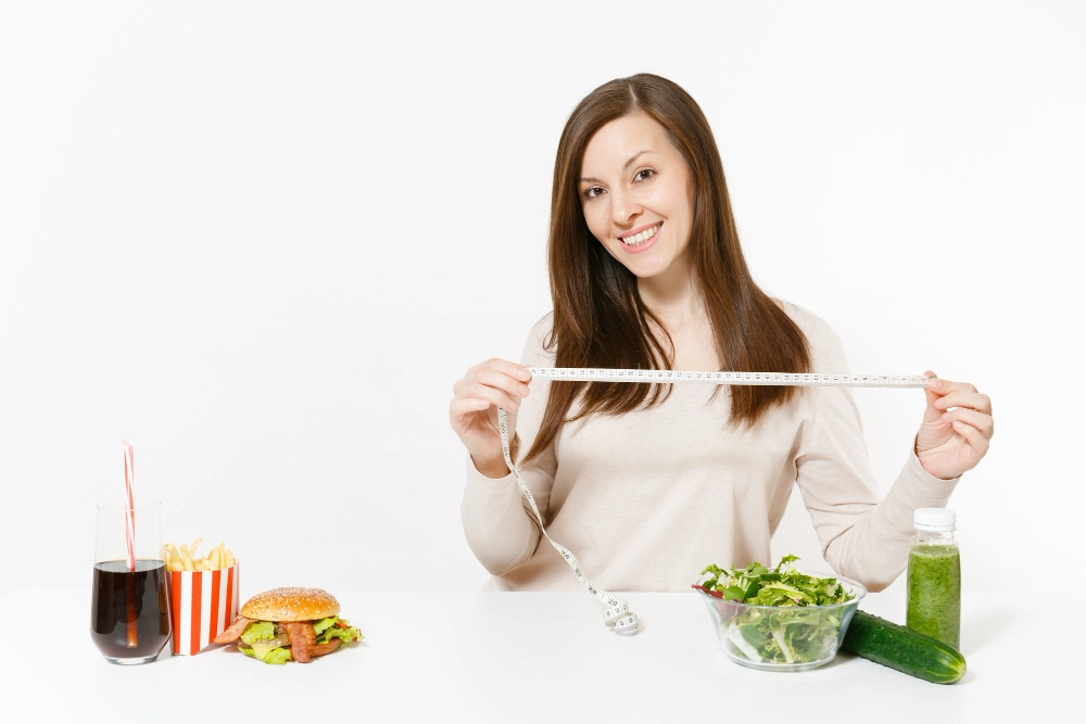 Woman with measure tape, green detox smoothies, salad in glass bowl, cucumber, burger, cola in bottle isolated on white background. Proper nutrition, healthy lifestyle, fast food, dieting concept.