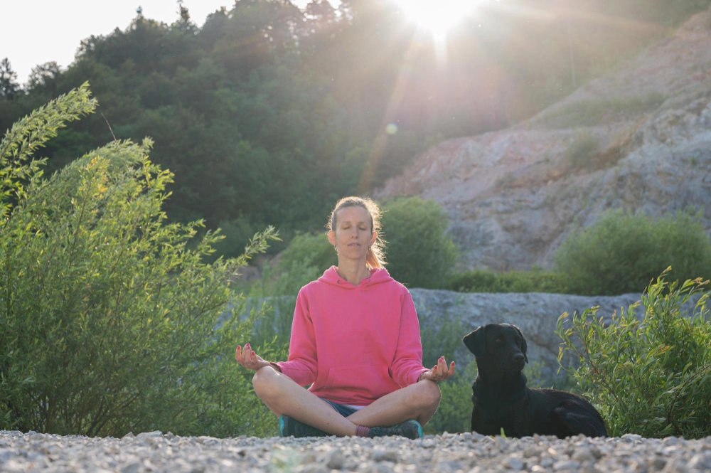 Young woman meditating in nature at sunrise with her dog beside her