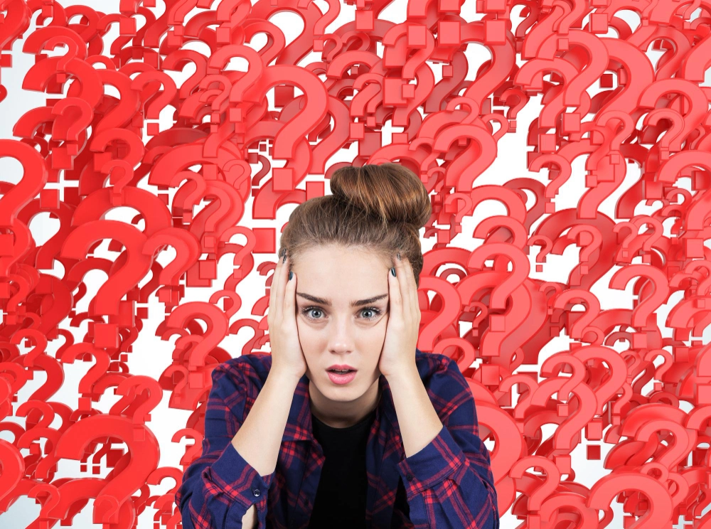 Portrait of a stressed out teen girl sitting against red question marks background. Concept of multiple questions that need to be answered