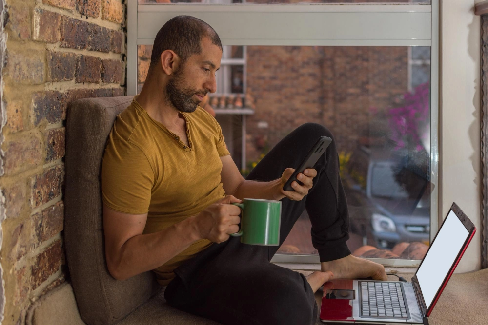 Young man with beard sweat pants and tshirt working at home having a hot drink while checking his smartphone